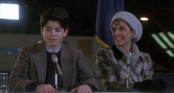Movie still from “Rocky V” (1990), directed by John G. Avildsen – A woman and a young boy sitting next to each other at microphones in front of an american flag; Medium shot, High angle