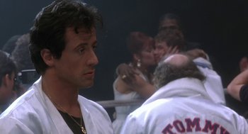 Movie still from “Rocky V” (1990), directed by John G. Avildsen – Sylvester stallone in a boxing ring with a group of people; Medium shot, Over the shoulder angle