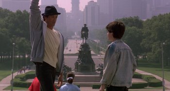 Movie still from “Rocky V” (1990), directed by John G. Avildsen – Two men standing in front of a statue in a city; Medium shot, Over the shoulder angle