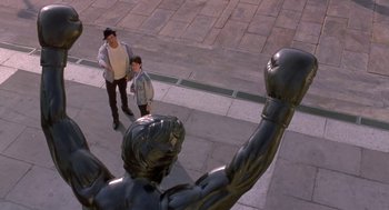 Movie still from “Rocky V” (1990), directed by John G. Avildsen – Two people standing in front of a statue of a man with a boxing glove; Wide shot, High angle