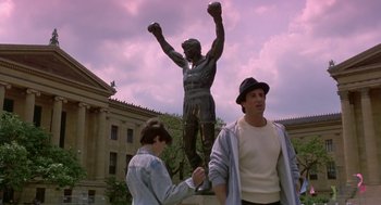 Movie still from “Rocky V” (1990), directed by John G. Avildsen – Two young men standing in front of a bronze statue of a man with boxing gloves; Wide shot, Low angle