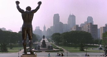 Movie still from “Rocky V” (1990), directed by John G. Avildsen – A view of a statue of a man in a park with a city in the background; Extreme Wide shot, Low angle