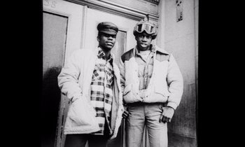 Movie still from “She's Gotta Have It” (1986), directed by Spike Lee – Two men standing next to each other in front of a building; Medium shot, Low angle