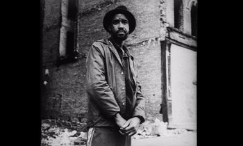 Movie still from “She's Gotta Have It” (1986), directed by Spike Lee – A black and white photo of a man standing in front of a building; Medium shot, Low angle