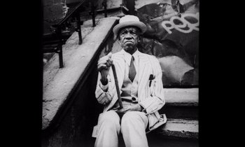 Movie still from “She's Gotta Have It” (1986), directed by Spike Lee – An older man sitting on the steps of a building; Medium shot, Low angle