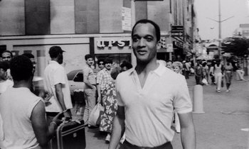Movie still from “She's Gotta Have It” (1986), directed by Spike Lee – Black and white photograph of a man walking in a crowded street; Medium shot, Low angle