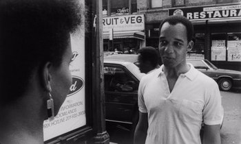 Movie still from “She's Gotta Have It” (1986), directed by Spike Lee – A black and white photo of a man standing on a street corner; Medium shot, Over the shoulder angle