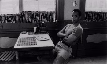 Movie still from “She's Gotta Have It” (1986), directed by Spike Lee – Black and white photograph of a man sitting at a table; Medium shot, High angle