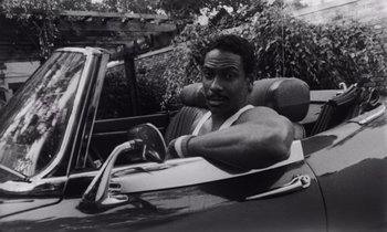Movie still from “She's Gotta Have It” (1986), directed by Spike Lee – A black and white photo of a man driving a car; Close Up shot, Low angle