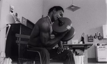 Movie still from “She's Gotta Have It” (1986), directed by Spike Lee – A black and white photo of a man lifting a weight; Medium shot, Low angle