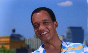 Movie still from “She's Gotta Have It” (1986), directed by Spike Lee – A man smiling for the camera with a city skyline in the background; Close Up shot, Low angle