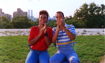 Movie still from “She's Gotta Have It” (1986), directed by Spike Lee – A man and a woman sitting on the grass clapping; Medium shot, Low angle