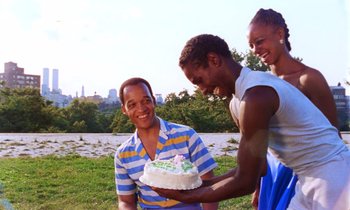 Movie still from “She's Gotta Have It” (1986), directed by Spike Lee – A man is holding a cake while two other people look on; Medium shot, Over the shoulder angle