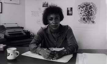 Movie still from “She's Gotta Have It” (1986), directed by Spike Lee – A woman sitting at a table with a pen in her hand; Medium shot, Low angle