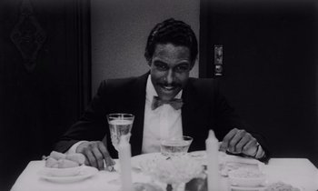 Movie still from “She's Gotta Have It” (1986), directed by Spike Lee – A man sitting at a dinner table smiling for the camera; Medium shot, High angle