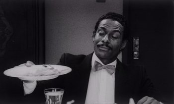 Movie still from “She's Gotta Have It” (1986), directed by Spike Lee – A black and white photo of a man sitting at a dinner table; Close Up shot, High angle