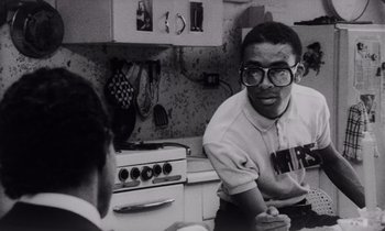 Movie still from “She's Gotta Have It” (1986), directed by Spike Lee – A young man wearing glasses is talking to another man in front of an oven in a kitchen; Medium shot, Over the shoulder angle
