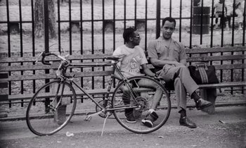 Movie still from “She's Gotta Have It” (1986), directed by Spike Lee – A man sitting on a bench next to a bicycle; Medium shot, Low angle
