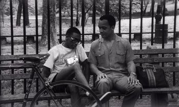 Movie still from “She's Gotta Have It” (1986), directed by Spike Lee – A man sitting on a bench next to another man with a bicycle; Medium shot, High angle