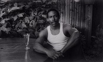 Movie still from “She's Gotta Have It” (1986), directed by Spike Lee – A black and white photo of a man sitting next to a glass of water; Medium shot, Low angle