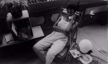 Movie still from “She's Gotta Have It” (1986), directed by Spike Lee – Black and white photograph of a woman sitting in a chair; Medium shot, High angle
