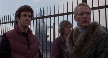 Movie still from “Slap Shot” (1977), directed by George Roy Hill – Two men standing next to each other in front of a metal fence; Medium shot, Low angle