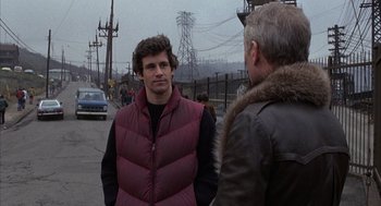 Movie still from “Slap Shot” (1977), directed by George Roy Hill – A man wearing a red vest talking to another man on the side of the road; Medium shot, Over the shoulder angle