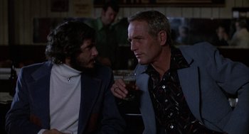 Movie still from “Slap Shot” (1977), directed by George Roy Hill – Two men sitting at a table with a glass of beer; Medium shot, Over the shoulder angle