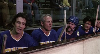 Movie still from “Slap Shot” (1977), directed by George Roy Hill – A group of men sitting next to each other on a hockey rink; Medium shot, High angle