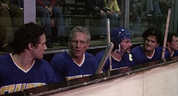 Movie still from “Slap Shot” (1977), directed by George Roy Hill – A group of men sitting next to each other on a hockey rink; Medium shot, High angle