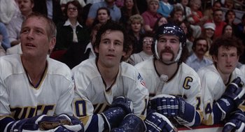 Movie still from “Slap Shot” (1977), directed by George Roy Hill – A group of hockey players sitting in front of an audience; Medium shot, Low angle