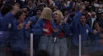 Movie still from “Slap Shot” (1977), directed by George Roy Hill – A couple of women standing next to each other in front of a group of people; Wide shot, Low angle