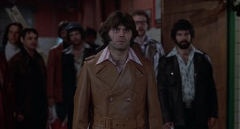 Movie still from “Slap Shot” (1977), directed by George Roy Hill – A group of people in a hallway with a man in a brown leather jacket; Medium shot, Low angle