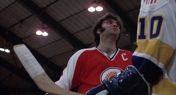 Movie still from “Slap Shot” (1977), directed by George Roy Hill – A man in a red and white hockey uniform holding a hockey stick; Close Up shot, Low angle