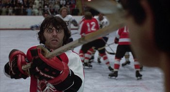 Movie still from “Slap Shot” (1977), directed by George Roy Hill – A man holding a baseball bat in front of a group of people; Close Up shot, Low angle