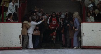 Movie still from “Slap Shot” (1977), directed by George Roy Hill – A crowd of people standing around a man in a red and white uniform; Wide shot, High angle