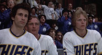 Movie still from “Slap Shot” (1977), directed by George Roy Hill – A group of men standing next to each other in front of an audience; Medium shot, Low angle