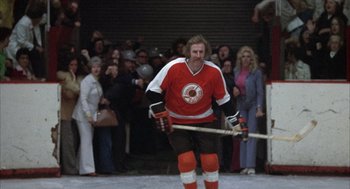 Movie still from “Slap Shot” (1977), directed by George Roy Hill – A man in a red and white hockey uniform holding a hockey stick; Wide shot, Low angle