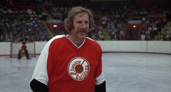 Movie still from “Slap Shot” (1977), directed by George Roy Hill – An older man in a hockey uniform stands in front of a crowd of people; Medium shot, Low angle