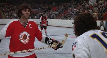 Movie still from “Slap Shot” (1977), directed by George Roy Hill – A man in a red and white jersey holding a hockey stick; Medium shot, Low angle