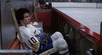 Movie still from “Slap Shot” (1977), directed by George Roy Hill – A man sitting on a bench in a hockey uniform; Medium shot, High angle