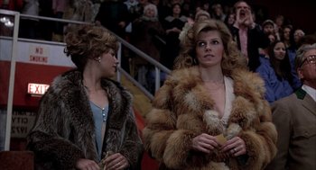 Movie still from “Slap Shot” (1977), directed by George Roy Hill – Two women in fur coats standing next to each other in front of a crowd of onlookers; Medium shot, Over the shoulder angle