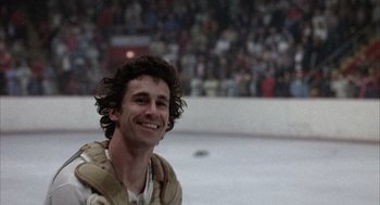 Movie still from “Slap Shot” (1977), directed by George Roy Hill – A man smiling at a hockey game while people watch from the stands; Close Up shot, Low angle