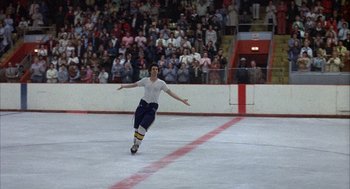 Movie still from “Slap Shot” (1977), directed by George Roy Hill – A man is skating on the ice in front of a crowd; Wide shot, Low angle