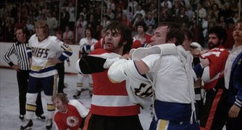 Movie still from “Slap Shot” (1977), directed by George Roy Hill – A hockey player in a red and white uniform is fighting with a player in a blue and white uniform; Medium shot, High angle