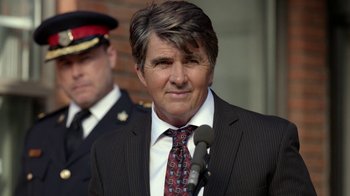 Movie still from “Slasher” (2016), created by Aaron Martin – A man in a suit and tie standing next to a microphone; Close Up shot, Over the shoulder angle