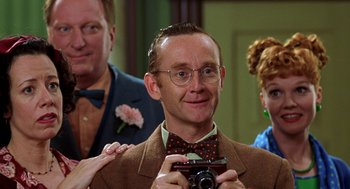 Movie still from “Stuart Little” (1999), directed by Rob Minkoff – A man in a suit and tie holding a camera in front of other people; Close Up shot, Over the shoulder angle