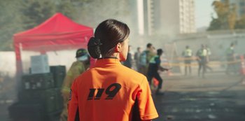 Movie still from “Sweet Home” (2020), directed by Eung Bok Lee – A woman in an orange shirt standing in front of a crowd of onlookers; Medium shot, Over the shoulder angle