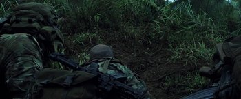 A man in a camouflage jacket and helmet is crouching in the grass