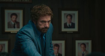 Movie still from “Air” (2023), directed by Ben Affleck – A man in a blue suit sitting in front of a wall of pictures; Medium shot, Over the shoulder angle